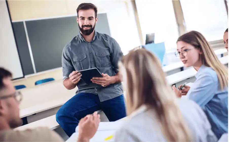 Ein Mann sitzt auf einem Tisch in einem Klassenzimmer mit einer Tafel an der Wand. Er hält ein iPad in der Hand und im Vordergrund diskutieren vier jüngere Personen miteinander