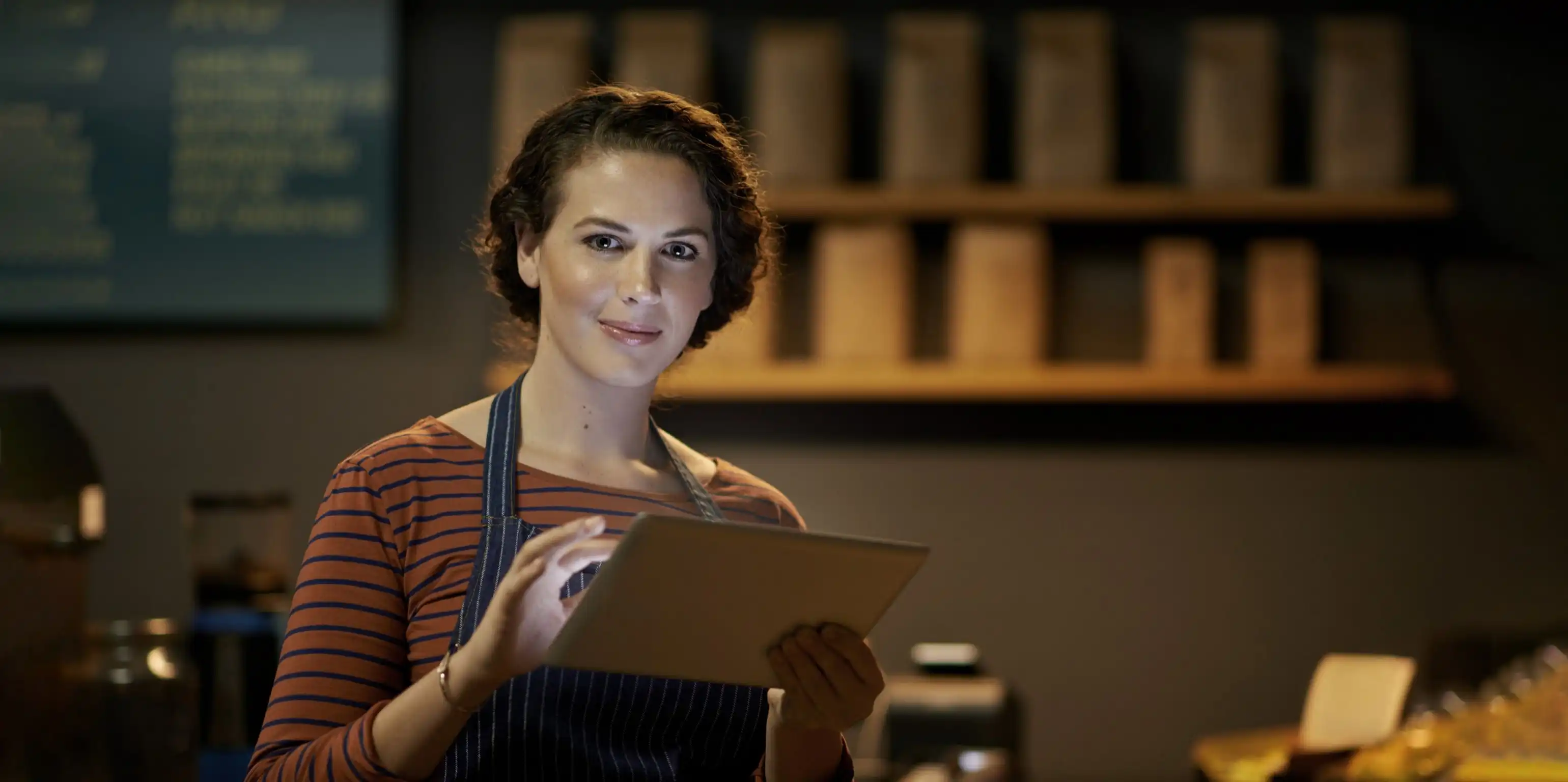 A woman wearing an apron is standing behind the counter of a café, holding a tablet