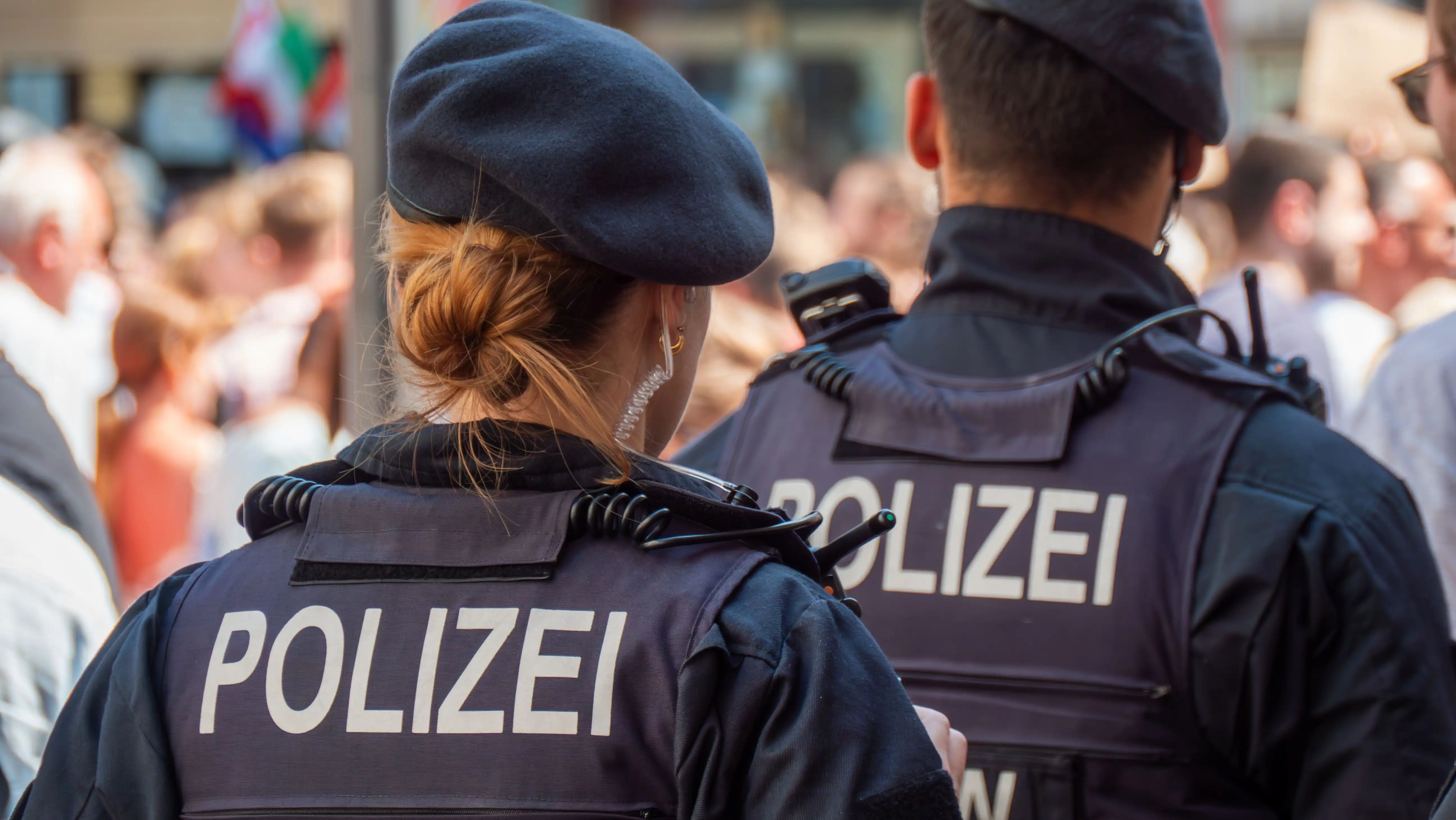 Zwei Polizeibeamte von hinten mit Schutzwesten und der Aufschrift ‚POLIZEI' blicken auf eine Menschenmenge.