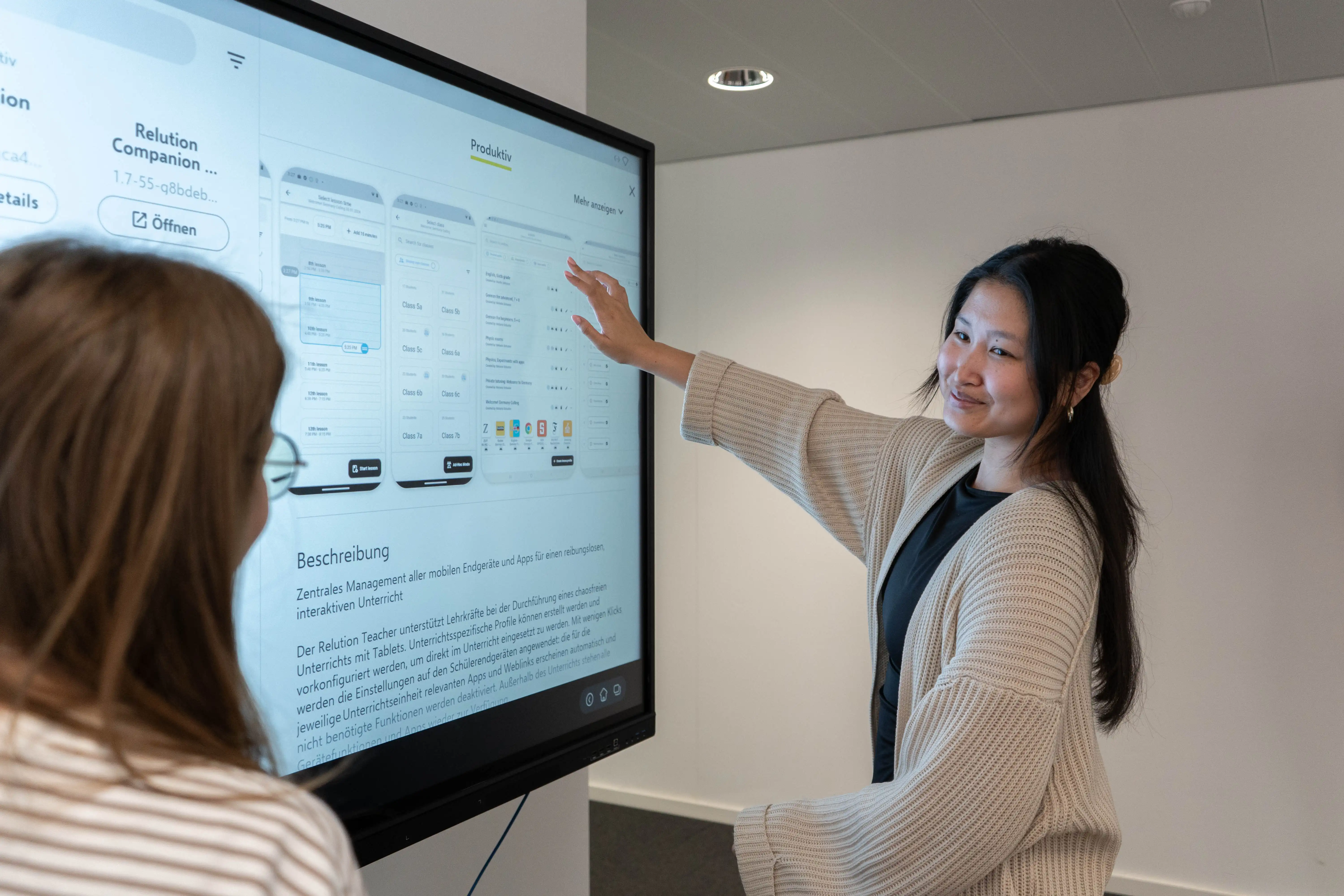 Eine Frau zeigt mit ihrem rechten Arm auf eine Grafik auf einem interaktiven Whiteboard, während sie zu einer zweiten Person blickt, die sie beobachtet