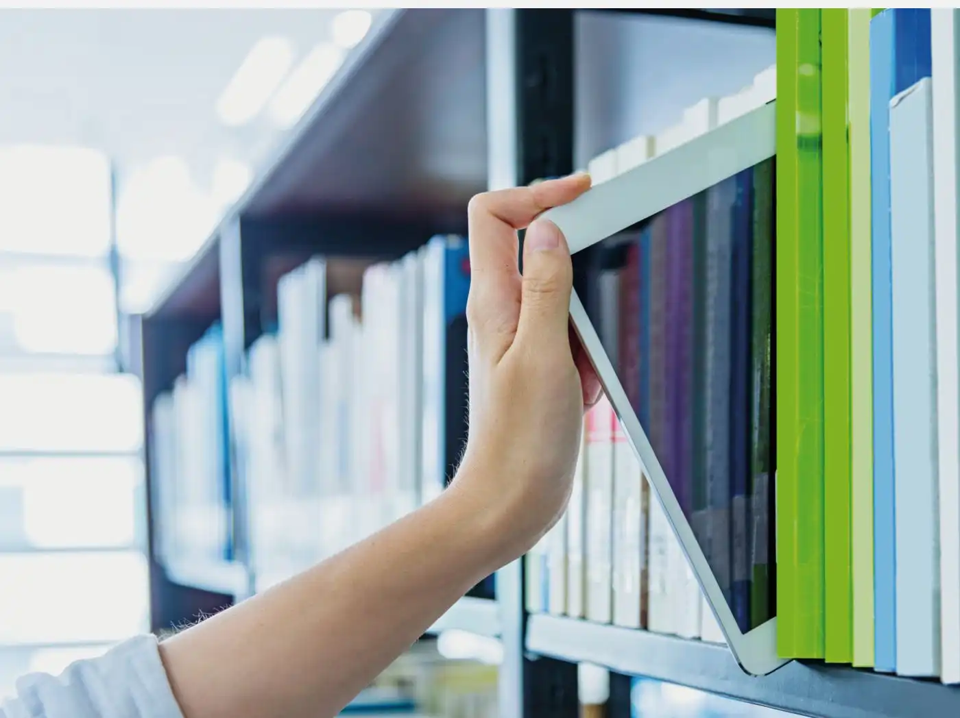 Eine Hand greift nach einem Tablet, welches zwischen zahlreichen Büchern in einem Bücherregal eingeordnet ist, im Hintergrund sind viele weitere Regale mit Büchern sichtbar