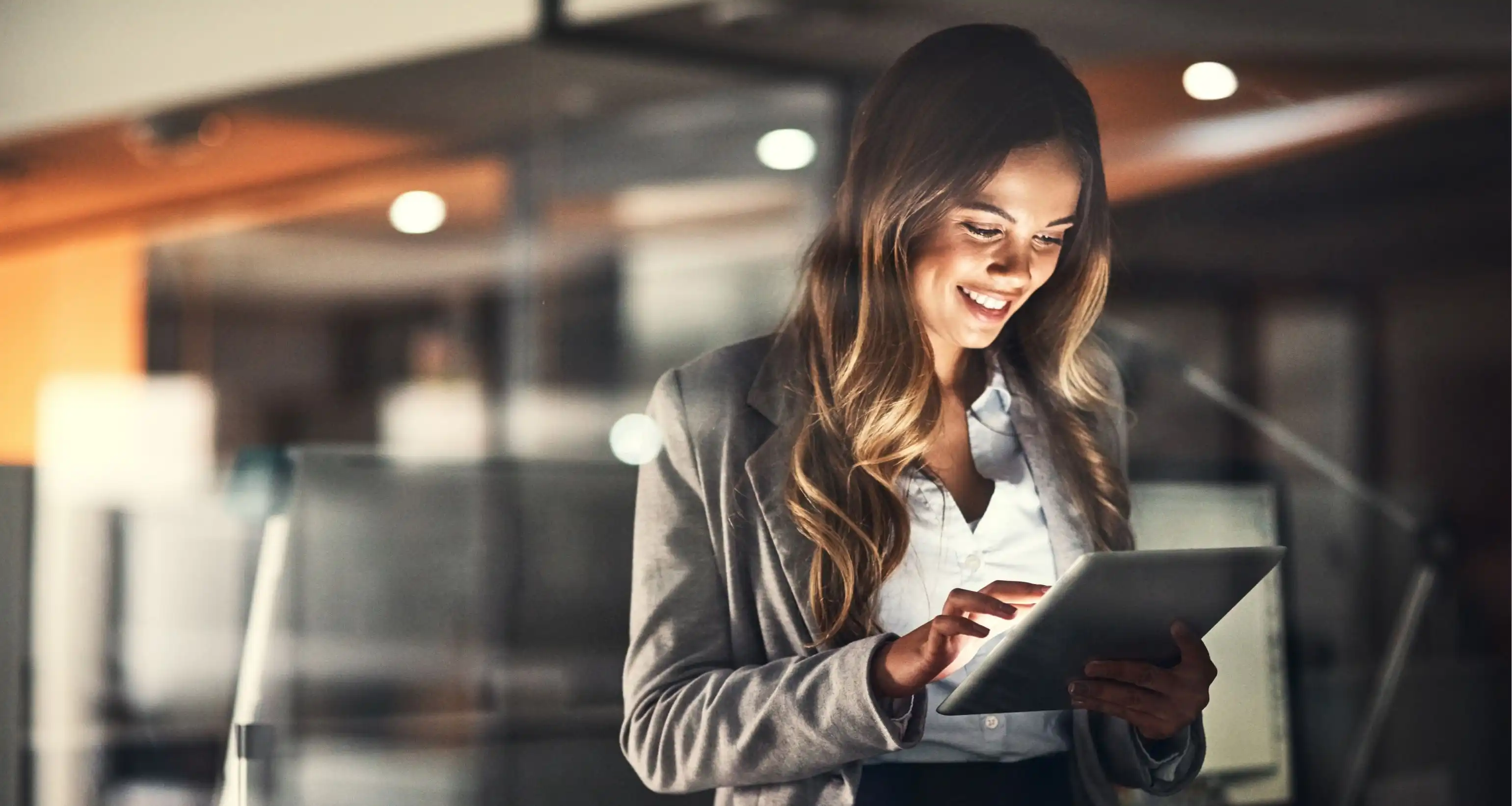 Eine Frau in Business-Kleidung hält ein Tablet in ihrer Hand, auf das sie lächelnd blickt, im Hintergrund ist ein modernes Büro sichtbar.