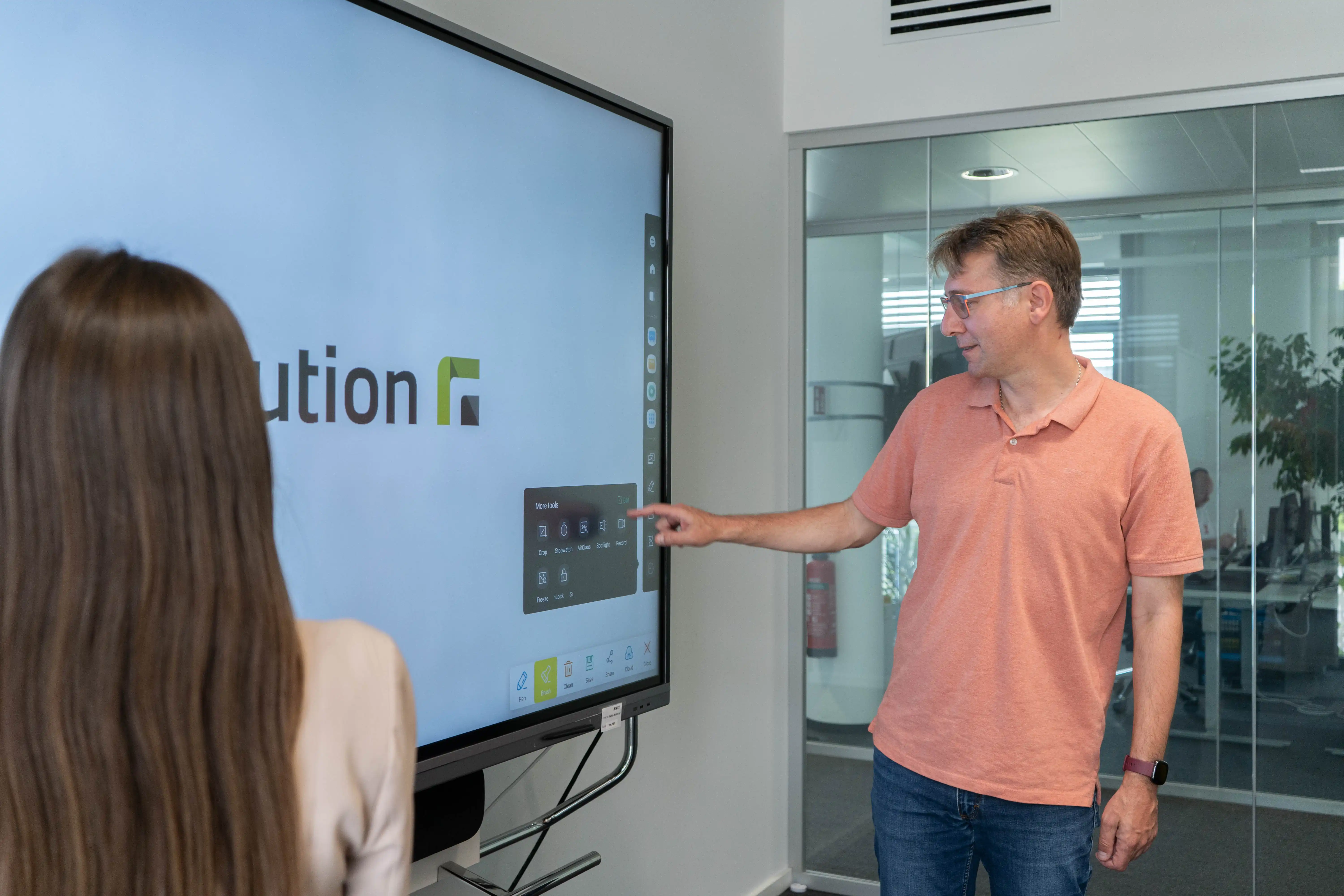 Ein Mann nutzt den Touchbildschirm eines Whiteboards um Einstellungen in einem Menü vorzunehmen, eine weitere Person, die mit dem Rücken zur Kamera steht, betrachtet ihn dabei