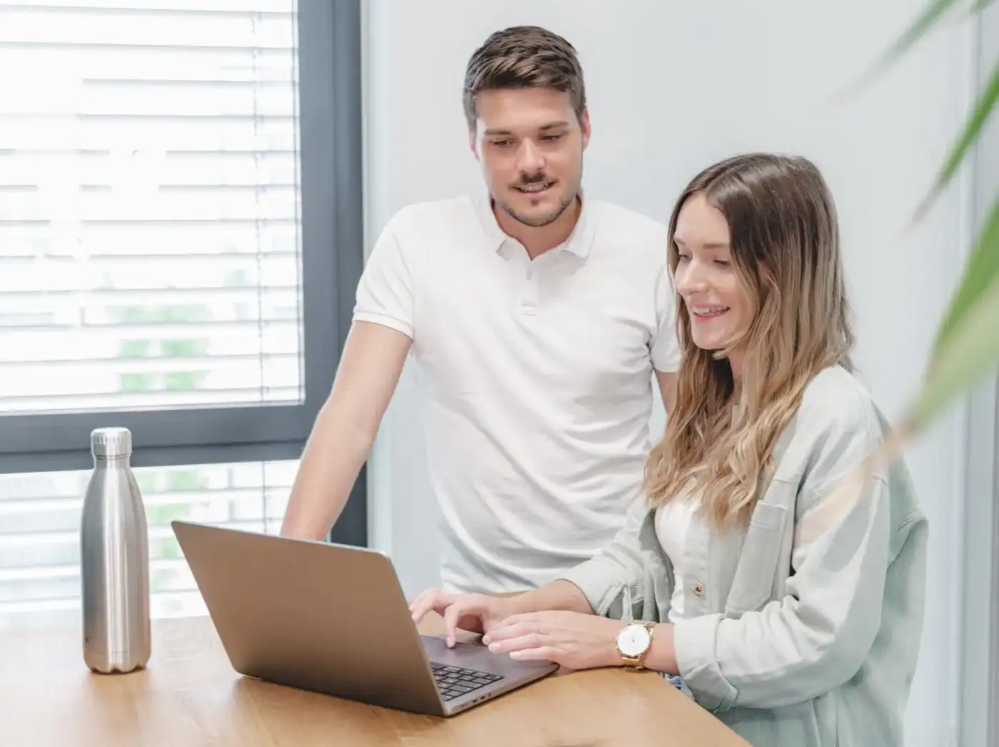 A man and a woman are standing in front of a laptop, looking together at its screen, while the woman has her finger on the touchpad