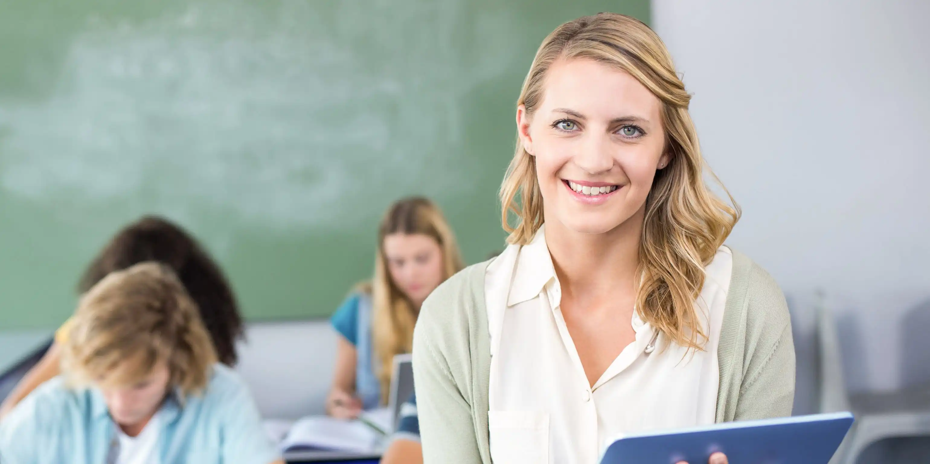 Eine Frau lächelt mit einem Tablet in der Hand in die Kamera. Hinter ihr sitzen drei jüngere Personen mit Heften und Stiften, im Hintergrund ist eine Schultafel sichtbar