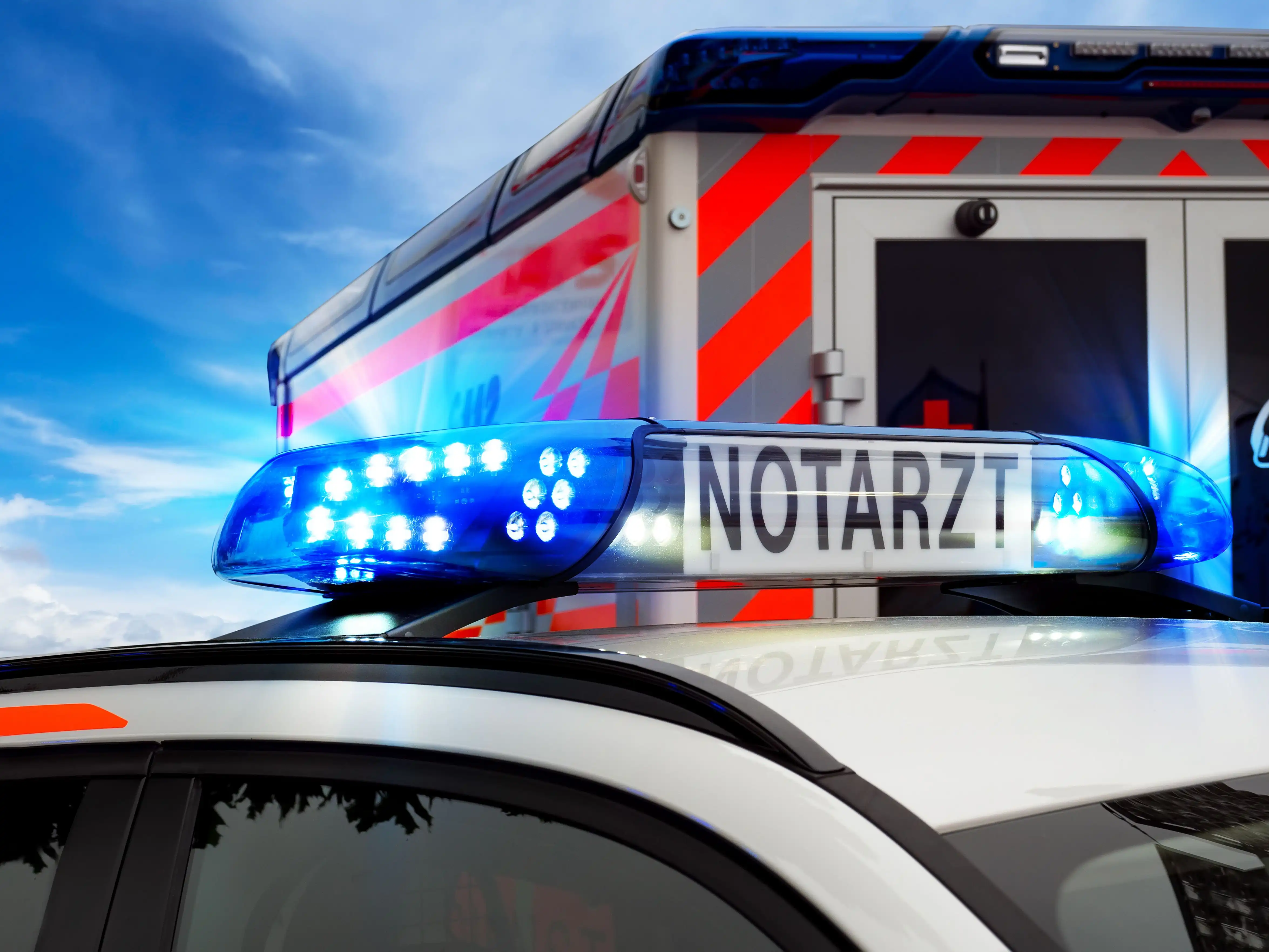 Leuchtender Blaulichtbalken mit der Aufschrift ‚NOTARZT' auf einem Fahrzeugdach, dahinter ein Rettungswagen vor blauem Himmel.