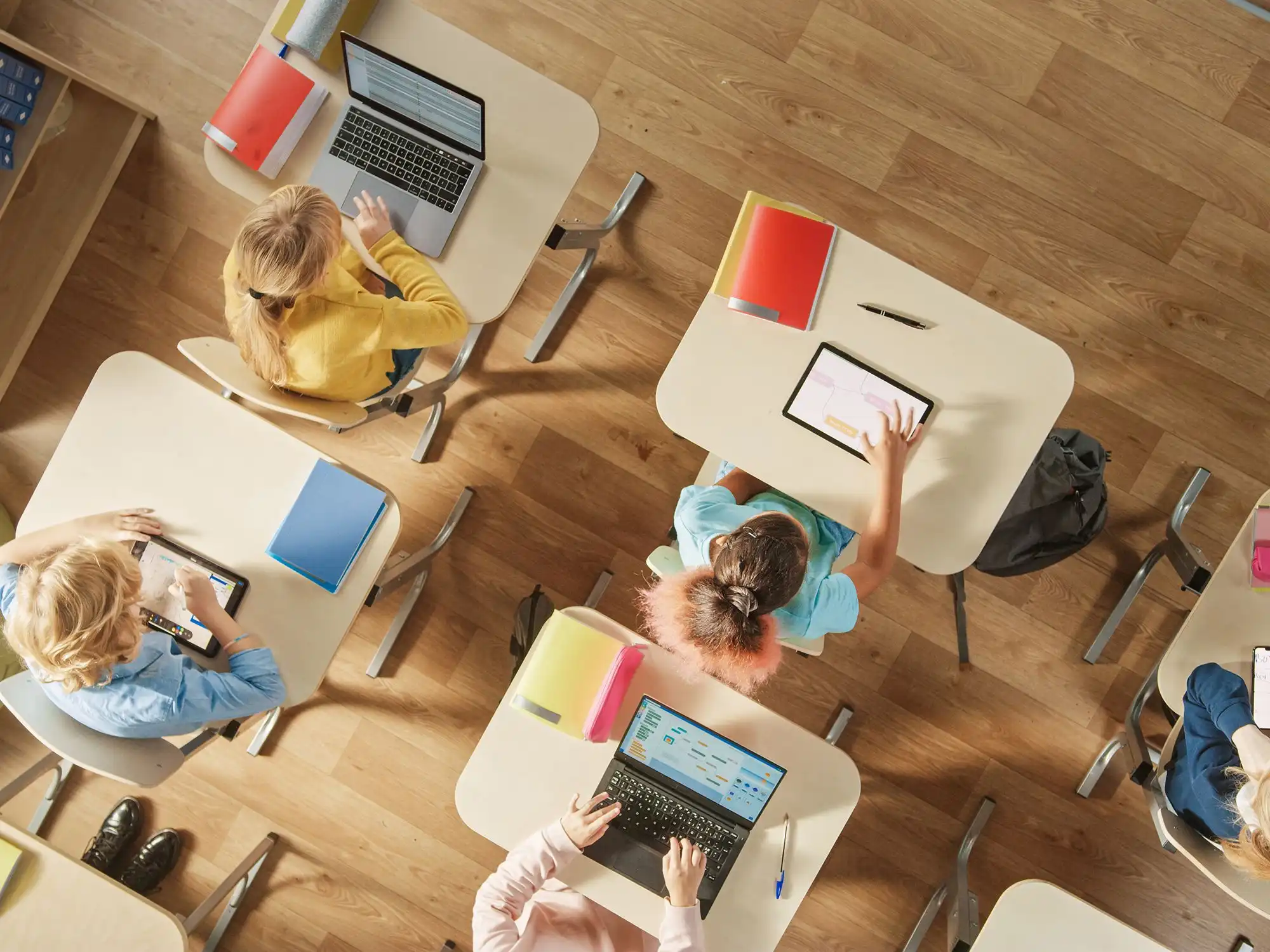 Vogelperspektive auf ein Klassenzimmer, in dem mehrere Kinder an Tischen mit Laptops und Tablets arbeiten
