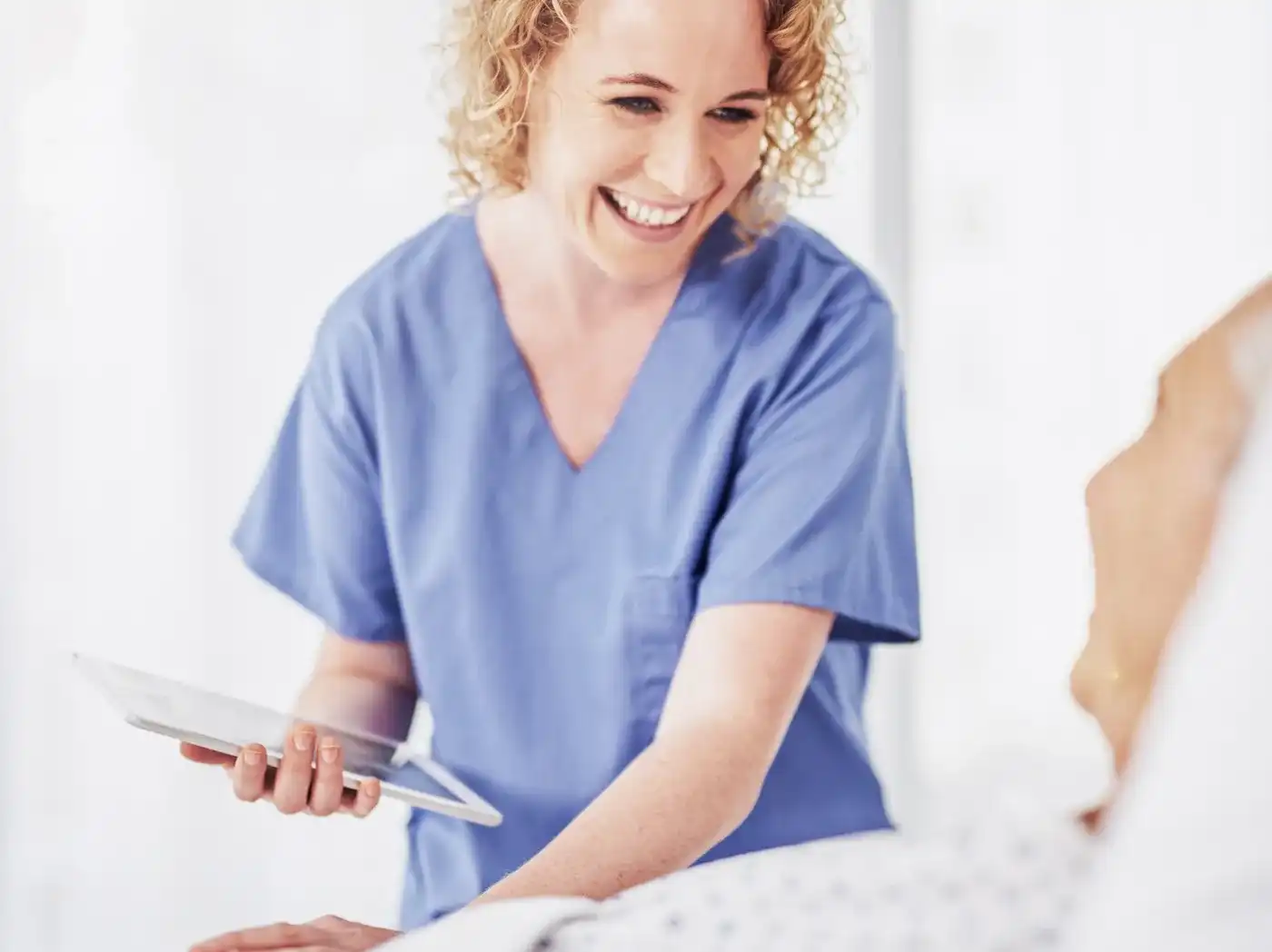 Eine lächelnde Frau in Krankenhauskleidung hält ein Tablet in ihrer Hand und kümmert sich um eine ältere Person in einem Bett