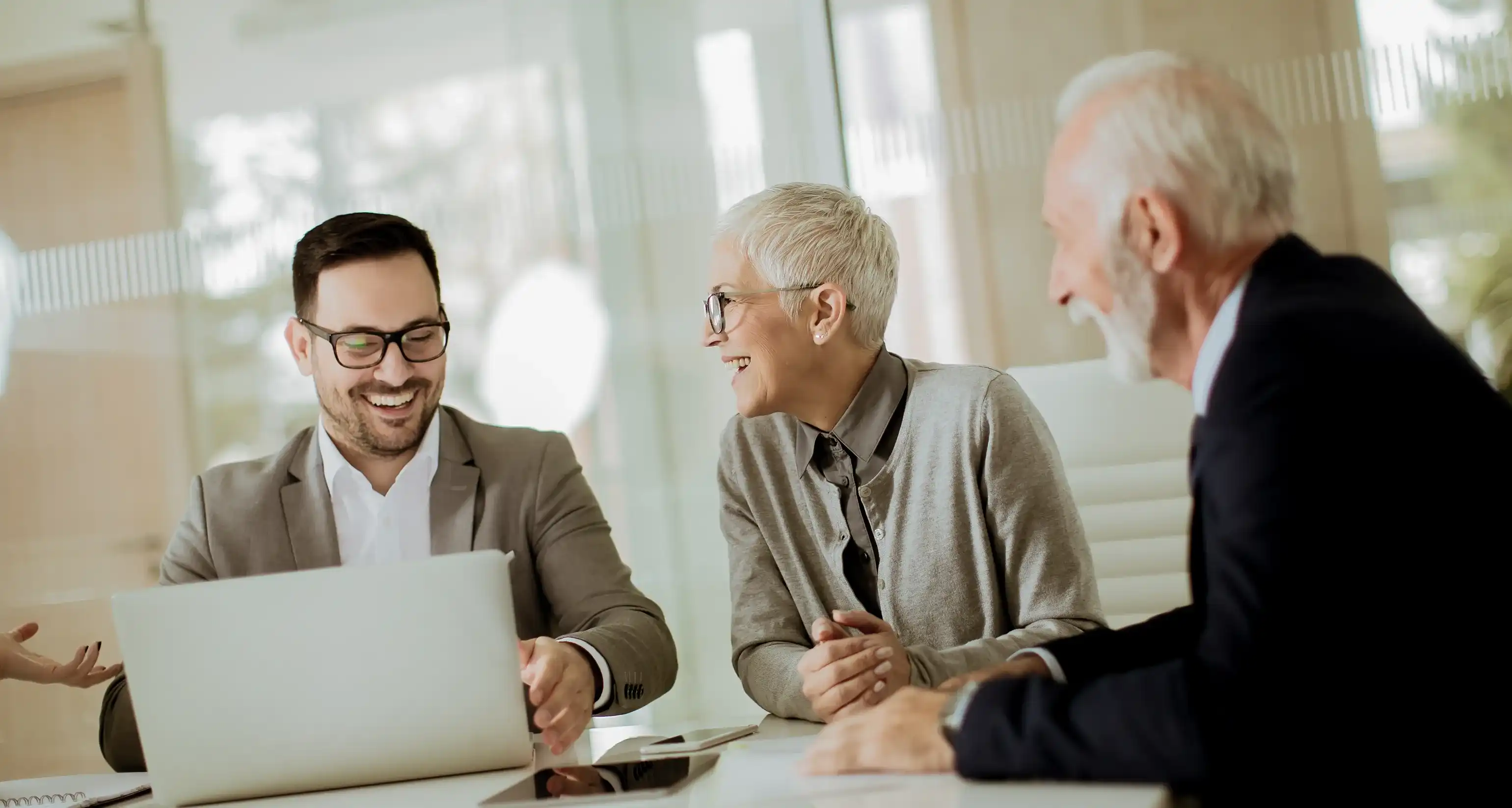 Ein jüngerer Mann sitzt an einem Tisch vor einem Laptop, neben ihm sitzen zwei ältere Personen, die ihn anschauen
