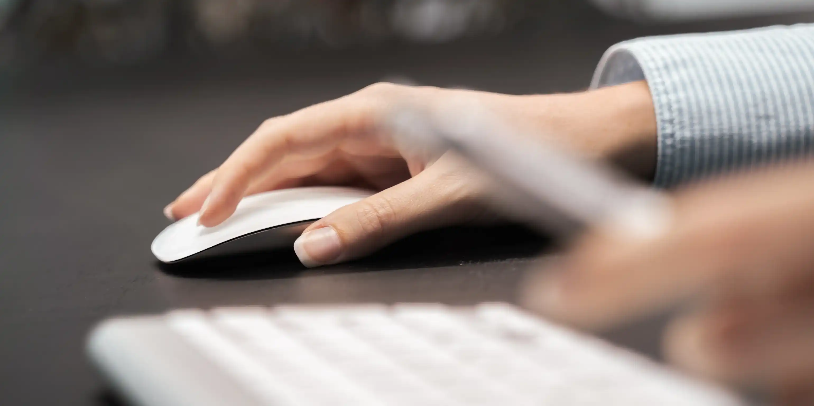 Auf dem Bild ist eine Hand sichtbar, die eine Computermaus nutzt, im Vordergrund sieht man eine Tastatur und unscharf eine weitere Hand