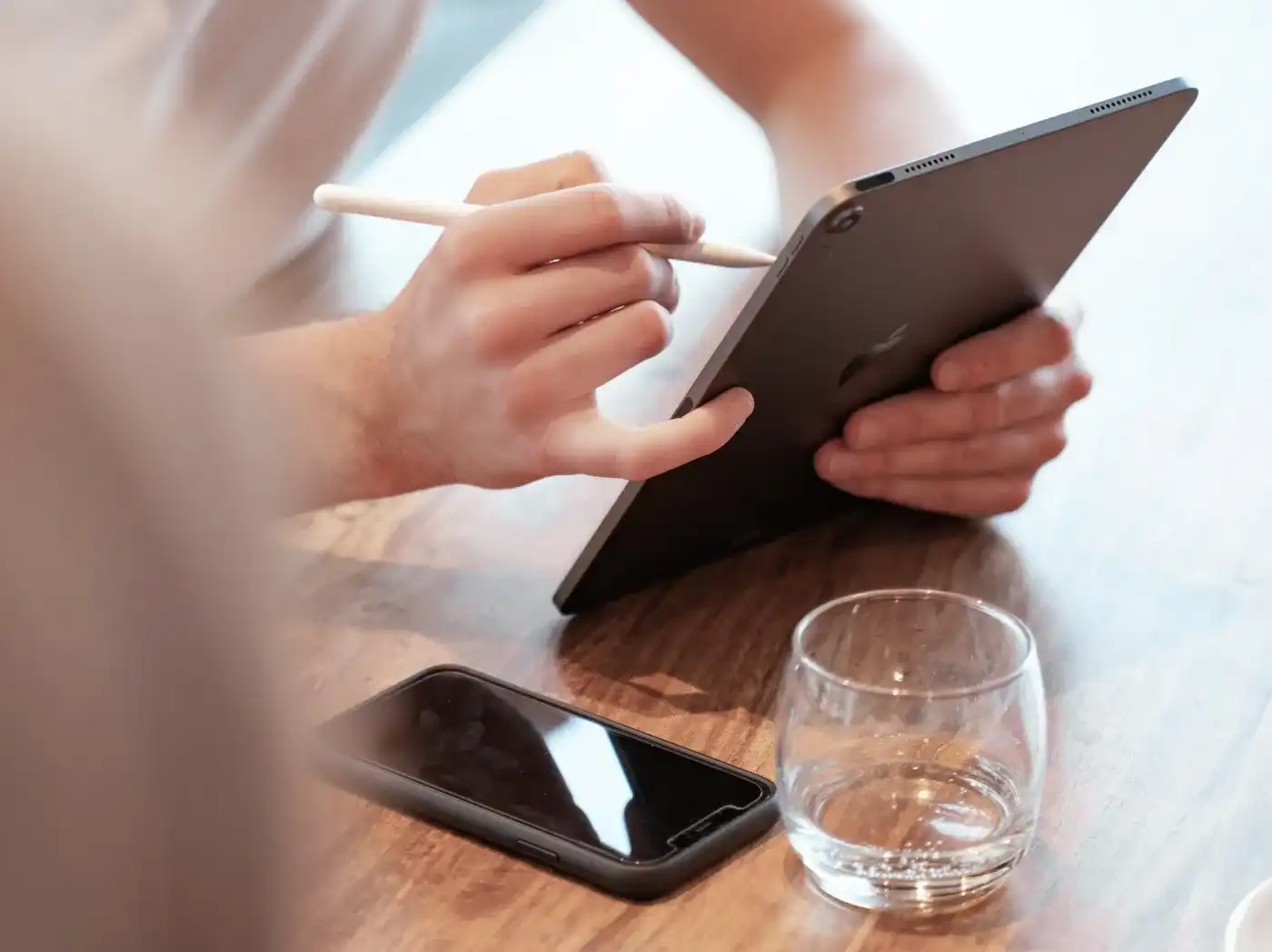 A person is working with an Apple Pencil on an iPad, with an iPhone lying next to them on the table