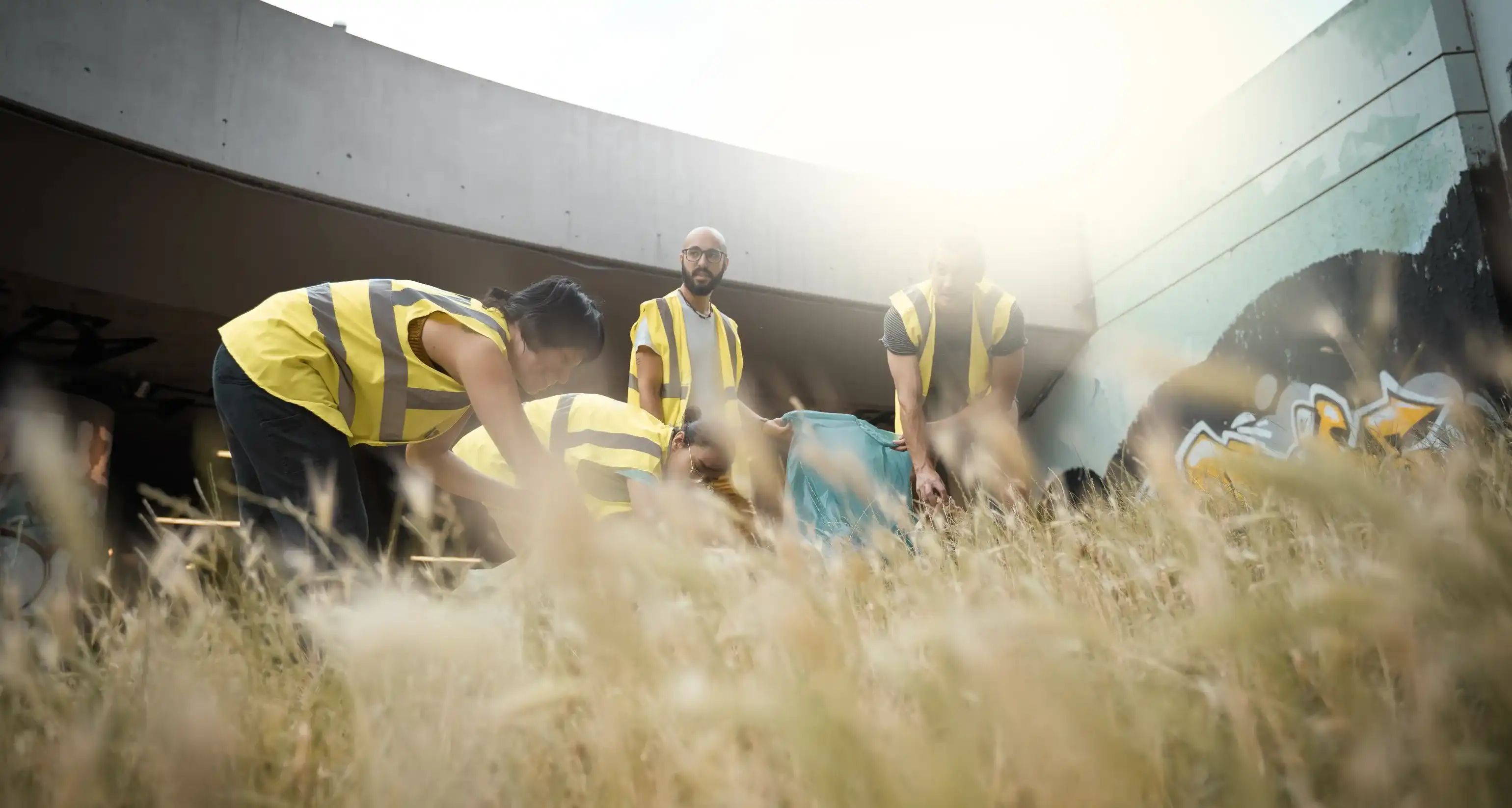 Vier Personen in gelben Warnwesten sammeln in einer Wiese, die sich zwischen Betongebäuden befindet, mit einer blauen Tüte Müll ein