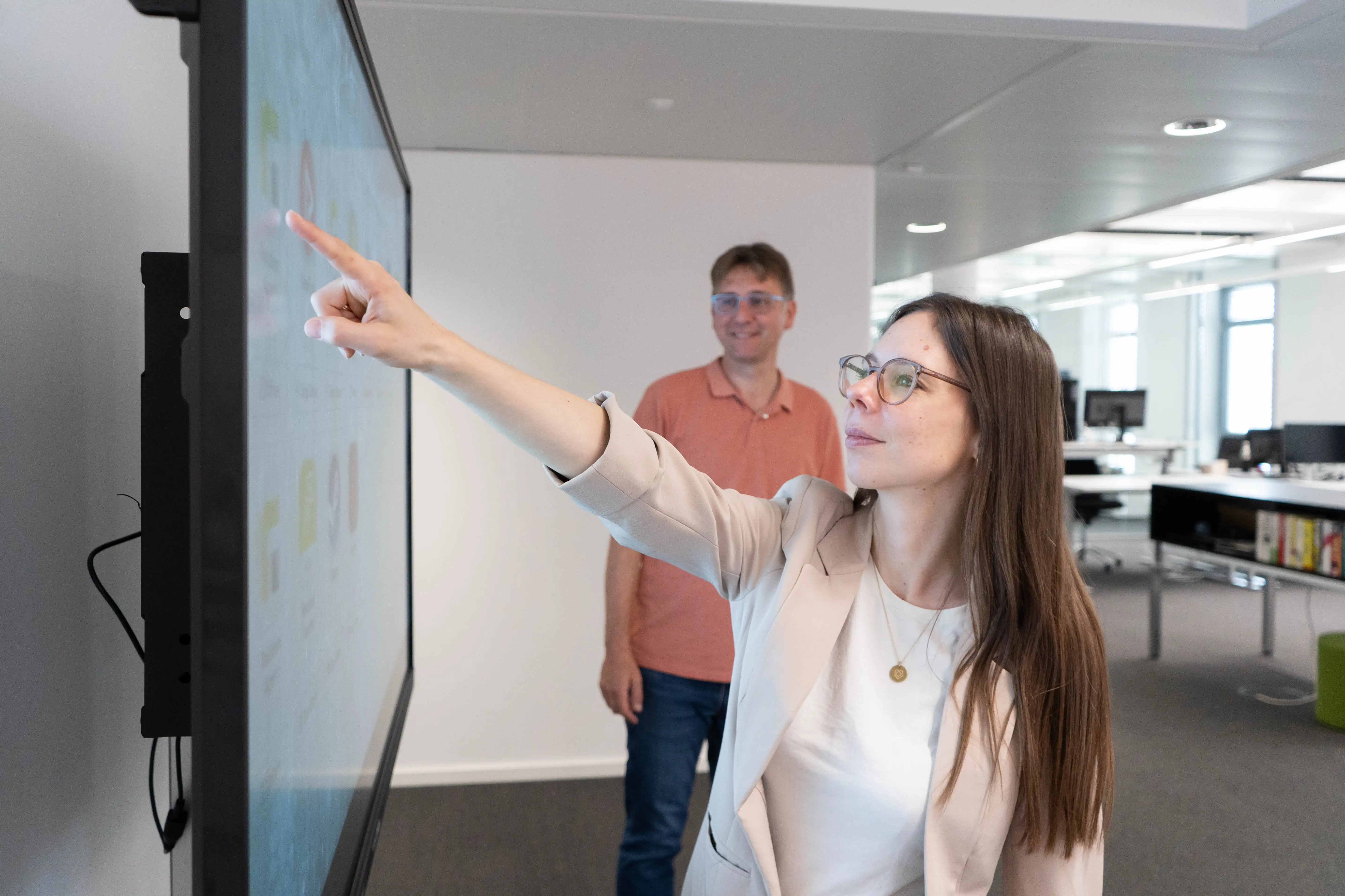 Eine Frau deutet mit dem Zeigefinger auf ein Symbol auf einem interaktiven Whiteboard, während ein lächelnder Mann etwas versetzt hinter ihr steht und beobachtet
