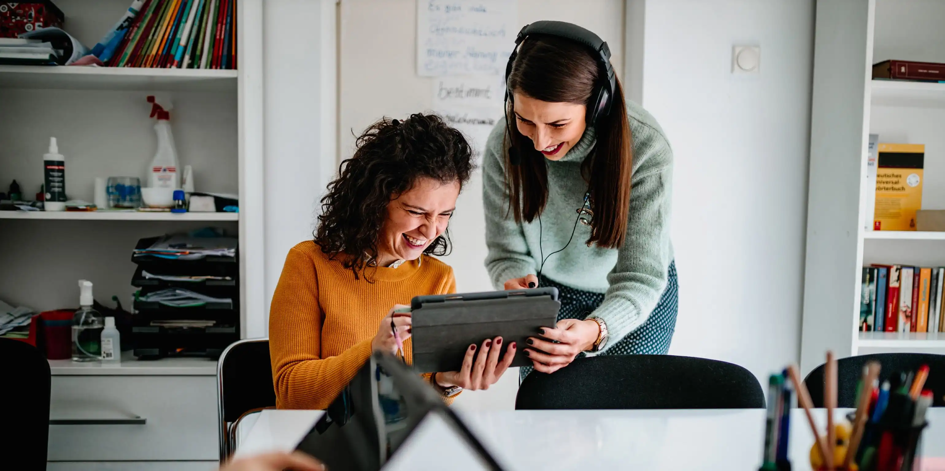 Zwei lachende Frauen halten gemeinsam ein Tablet in der Hand, eine der beiden hat Kopfhörer auf, die mit dem iPad verbunden sind, beide befinden sich in einem Raum mit zahlreichen Lernutensilien